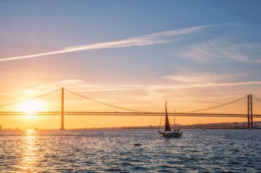 Lizbon 'un ünlü turistik beldesi 25 de Abril Köprüsü' nün Tagus nehri üzerinde Lisboa ve Almada 'yı gün batımında turistik yat siluetiyle bağlayan görüntüsü. Lizbon, Portekiz