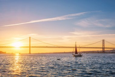 Lizbon 'un ünlü turistik beldesi 25 de Abril Köprüsü' nün Tagus nehri üzerinde Lisboa ve Almada 'yı gün batımında turistik yat siluetiyle bağlayan görüntüsü. Lizbon, Portekiz