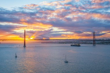 25 de Abril Köprüsü 'nün Tagus Nehri üzerinde gün batımında turistik yat tekneleri ve kargo konteynır gemisiyle Lizbon' un ünlü turistik tarihi manzarası. Lizbon, Portekiz