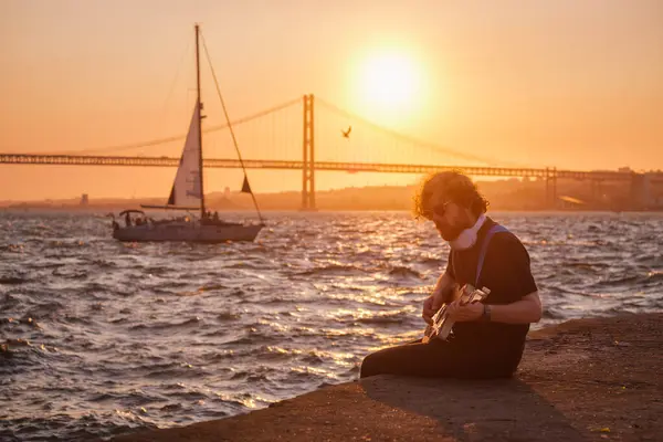 希波斯特街头音乐家穿着黑色的电吉他 在日落时分在街上的堤岸上弹奏 背景是四月二十五日的桥和游艇 葡萄牙里斯本 — 图库照片