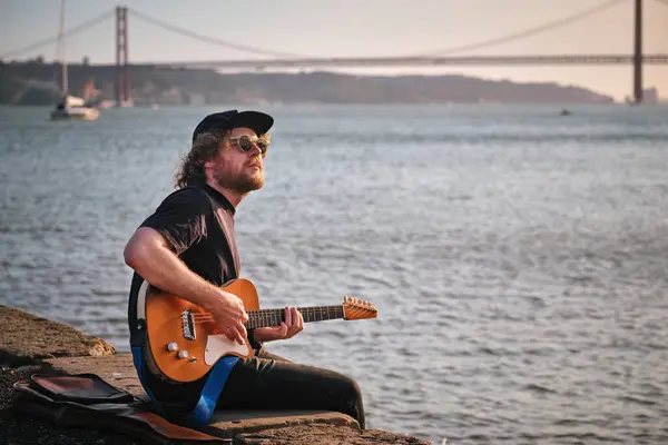 Hipster street musician in black playing electric guitar in the street sitting on pier embankment on sunset