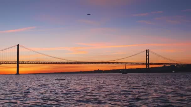 リスボンとアルマダを結ぶリスボンの有名な観光ランドマークである25 Abril Bridgeの眺め タガス川の上のセトゥーバル半島にあり 夕日と飛行機のヨットのシルエット ポルトガルのリスボン — ストック動画