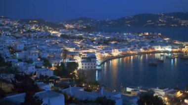 Meşhur yel değirmenleriyle Mykonos kasabası manzarası, ve gece mavi saatte aydınlatılmış tekneleri ve yolcu gemisiyle liman manzarası. Mykonos, Cyclades adaları, Yunanistan. Kamera kaydıyla
