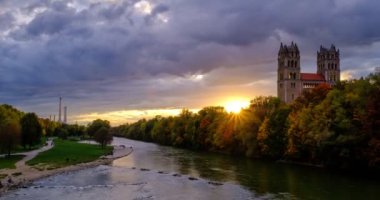 Münih 'in dramatik günbatımında geçen zamanı: Bavyera' nın ünlü turistik kentinde sonbahar. Reichenbach Köprüsü 'nden Isar nehri, ağaçlar ve kilise manzarası. Munchen, Bavyera, Almanya. Kamera yakınlaştır