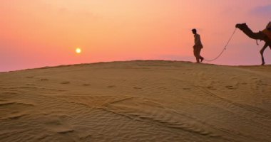 Gün batımında Thar Çölü 'nün kum tepelerindeki deve siluetleriyle iki Kızılderili kambur (deve sürücüsü). Rajasthan 'daki karavan turizm arka planında safari macerası. Jaisalmer, Rajasthan, Hindistan