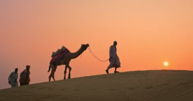 Gün batımında Thar Çölü 'nün kum tepelerindeki deve siluetleriyle Hintli kambur (deve sürücüsü). Rajasthan 'daki karavan turizm arka planında safari macerası. Jaisalmer, Rajasthan, Hindistan