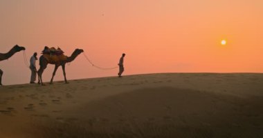 Gün batımında Thar Çölü 'nün kum tepelerindeki deve siluetleriyle iki Kızılderili kambur (deve sürücüsü). Rajasthan 'daki karavan turizm arka planında safari macerası. Jaisalmer, Rajasthan, Hindistan