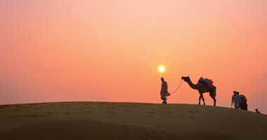 Gün batımında Thar Çölü 'nün kum tepelerindeki deve siluetleriyle Hintli kambur (deve sürücüsü). Rajasthan 'daki karavan turizm arka planında safari macerası. Jaisalmer, Rajasthan, Hindistan