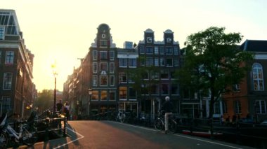 AMSTERDAM, NETHERLANDS - 20 Mayıs 2018: Amsterdam caddesinde gün batımında köprü ve ortaçağ evleriyle dolaşan yaya ve bisikletçiler