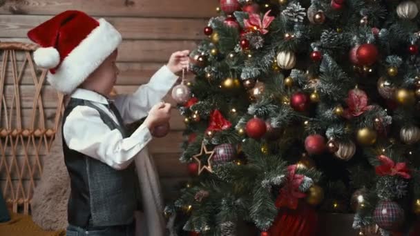 Kleiner Junge Mit Nikolausmütze Schmückt Weihnachtsbaum Mit Christbaumkugeln — Stockvideo