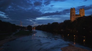 Münih doğası dramatik bir şekilde alacakaranlıkta: Turist Bavyera şehrinin parkında sonbahar. Isar Nehri manzarası, ağaçlar ve Reichenbach Köprüsü 'nden aydınlatılmış St. Maximilian kilisesi. Munchen, Bavyera, Almanya.