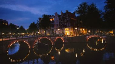 Amsterdam, Hollanda - 22 Mayıs 2018: Köprünün altından geçen tekneyle akşam alacakaranlığında Amsterdam şehir manzarası, kanal, köprü ve ortaçağ evleri aydınlatıldı