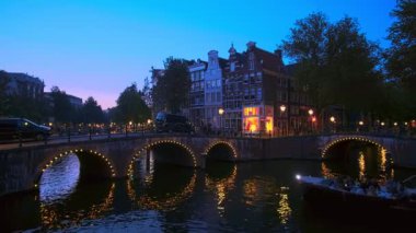 Amsterdam, Hollanda - 20 Mayıs 2018: Köprünün altından geçen tekneyle akşam alacakaranlığında Amsterdam şehir manzarası, kanal, köprü ve ortaçağ evleri aydınlatıldı