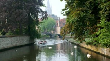 Bruges, Belçika - 29 Mayıs 2018: Bruges kanalı, arka planda Meryem Ana Kilisesi ve turist teknesinin geçtiği yaşlı ağaçların arasında su üzerinde beyaz kuğular. Brugge, Belçika