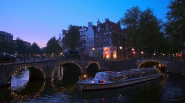 Amsterdam, Hollanda - 20 Mayıs 2018: Köprünün altından geçen turist tekneleriyle aydınlanan akşam alacakaranlıklarında Amsterdam 'ın kanal, köprü ve ortaçağ evlerinin gece manzarası