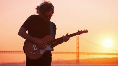 Günbatımı siluetinde sokakta elektro gitar çalan siyahlı hippi sokak müzisyeni.