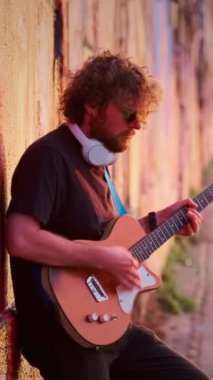 Günbatımında sokakta elektro gitar çalan siyahlı hippi sokak müzisyeni boyalı bir duvara yaslanıyor.