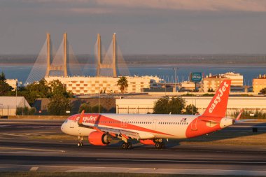 Lizbon, Portekiz - 11 Eylül 2023: easyJet Airbus A321-251NX yolcu uçağı taksisi Lizbon 'daki Humberto Delgado Havalimanı' nda günbatımında arka planda Vasco da Gamma köprüsü