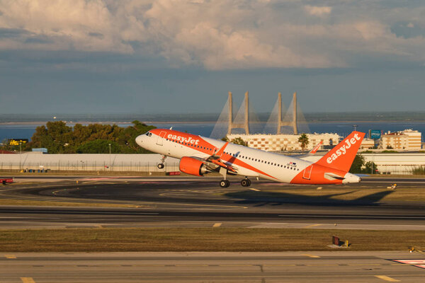 Лиссабон, Португалия - 11 сентября 2023 года: пассажирский самолет easyJet Airbus A320-251N взлетает в аэропорту Умберто Дельгадо в Лиссабоне на закате с мостом Васко да Гамма на заднем плане