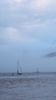 Lizbon 'un ünlü turistik beldesi 25 de Abril Köprüsü' nün Lisboa ve Almada 'yı yoğun sis bulutu altında, altından geçen teknelerle birbirine bağlayan görüntüsü. Lizbon, Portekiz