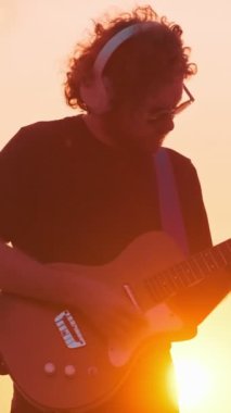 Günbatımı siluetinde sokakta elektro gitar çalan siyahlı hippi sokak müzisyeni.