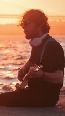 Hippi sokak müzisyeni, 25 Nisan köprüsü ve arka planda yatıyla günbatımında sokakta elektro gitar çalıyor. Lizbon, Portekiz