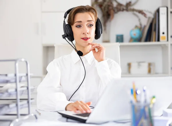 Portrait of young woman wearing headset, using laptop. Call center worker sitting at desk in office.