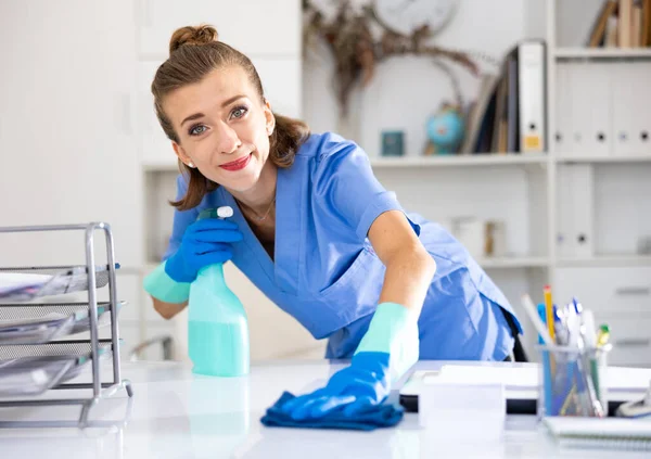 Young positive woman cleaner in overall polishing table in office with rag and detergent.