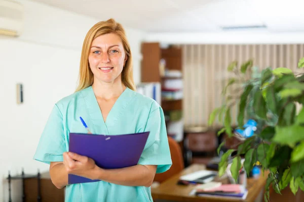 クリップボードでオフィスに立つ朗らかな笑顔の看護師の女性 医学史シートを書く — ストック写真