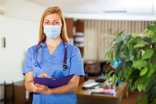 フレンドリーな女性医師の肖像マスク身に着けています制服スタンディングとともにStethoscopyで現代クリニック — ストック写真