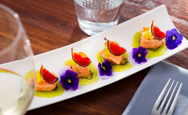 Pieces of pickled salmon served on kiwi slices with figs, mango and kumquat garnished with pansy flowers