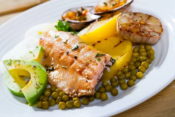 Delicious baked trout fillet served with vegetables, avocado and dog cockles on white plate