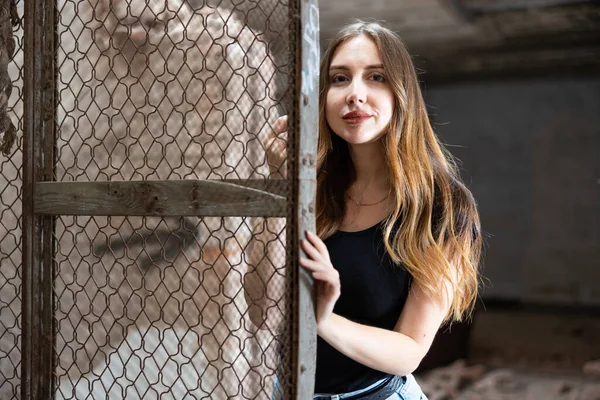 现代年轻的棕色头发的女人穿着黑色T恤和蓝色牛仔裤 站在铁丝网栅栏旁边 被废弃的大楼里 照片拍摄在愤怒的背景下 — 图库照片