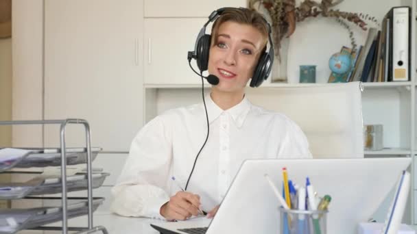 ノートパソコンを使用して ヘッドセットを身に着けている肯定的な若い女性の肖像 オフィスのデスクに座っているコールセンターの労働者 — ストック動画