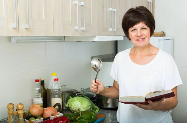 在家厨房里煮汤的快乐成熟女人检查菜谱的肖像 — 图库照片