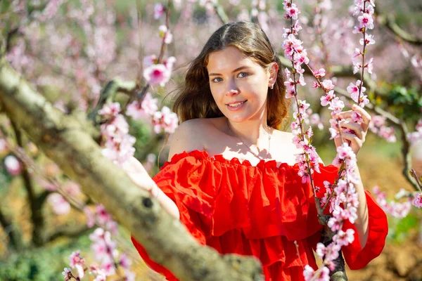 特写迷人的笑容可亲的女人 身穿红裙 站在一棵盛开的桃树背景下 — 图库照片