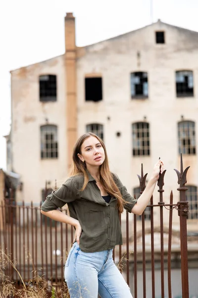 迷人的年轻女性 穿着蓝色牛仔裤和深色上衣 站在靠近金属栅栏的地方 背景是老式建筑 — 图库照片