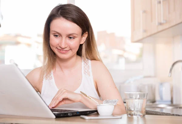 Smiling woman is working with laptop sitting at the table at her home.
