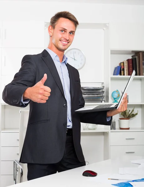 一个面带微笑的商人站在办公室手里拿着电脑的画像 — 图库照片