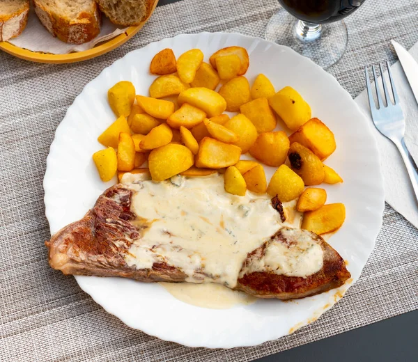 Portion of beefsteak with blue cheese sauce and potatoes served on plate with serving pieces.
