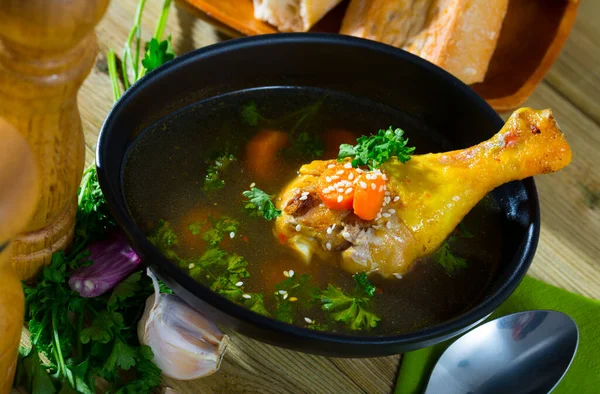 stock image Rustic style broth with chicken leg, vegetables and greens served in black bowl with fresh baguette