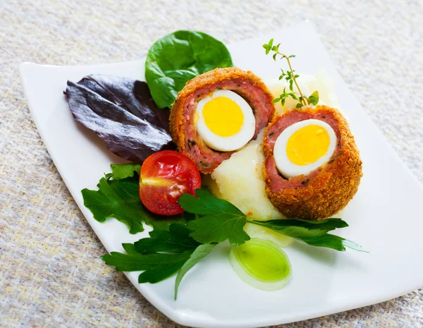 Halved Scotch eggs from quail eggs with garnish of mashed potatoes, vegetables and greens on white plate