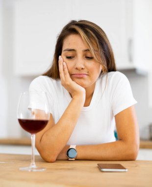 Upset young woman with glass of red wine in kitchen reading messages on mobile phone screen clipart