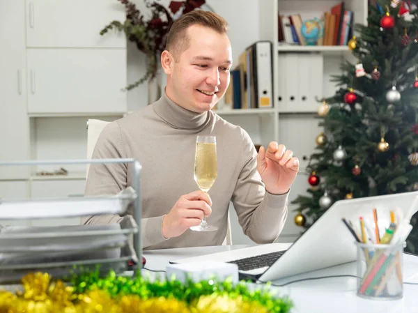 クリスマスの装飾されたオフィスのテーブルに座っている陽気な男のオフィスワーカーの肖像 シャンパンのガラスを保持し ビデオ通話中にジェスチャー — ストック写真