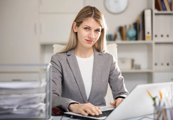 一个积极的微笑的女人坐在办公室的工作位置上 一边在笔记本电脑后面工作 — 图库照片