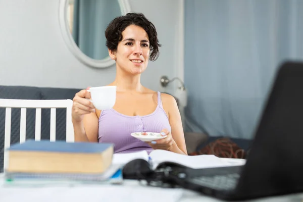 部屋のテーブルでノートパソコンの前でコーヒーを飲んで満足した女性を笑って — ストック写真