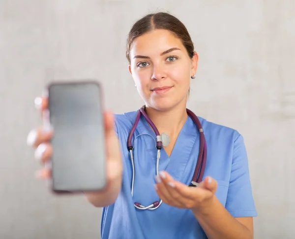 Młoda Uśmiechnięta Pielęgniarka Pokazująca Ekran Swojego Telefonu — Zdjęcie stockowe