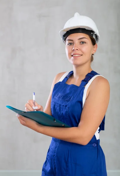 身穿蓝色工作服 头戴保护头盔的情绪化机修工女孩的画像 她在文档中写道 — 图库照片