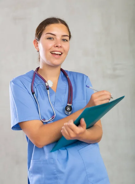 穿着蓝色制服的有礼貌的年轻女全科医生用病历填写剪贴板 灰色背景的演播室摄影 — 图库照片