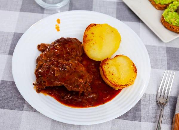 Delicious fried pork cheeks served in sauce with baked potatoes on plate